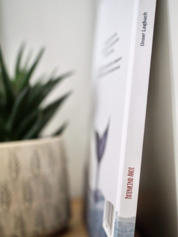 Das Erinnerungsbuch "Patenkind Ahoi" steht in einem Regal, auf dem Buchrücken steht "PATENKIND AHOI" und "Unser Logbuch"